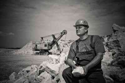 Black and white photo of a guy wearing a construction hat, sitting on a pile of rocks, in front of an old excavator. Sometimes increasing productivity means finding someone smarter than you to figure things out.